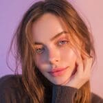 AI-generated portrait of a brunette woman with blue-grey eyes resting her hand on her cheek in a grey turtleneck sweater against a lavender to blush pink split gradient background for an Instagram reel cover