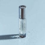 AI-generated product photo of a clear glass cylindrical skincare serum bottle with silver chrome cap surrounded by scattered water droplets on a cool light blue background