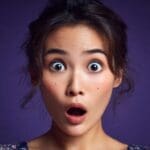 Front-facing portrait of a woman with dark hair pulled back in a shocked pose.